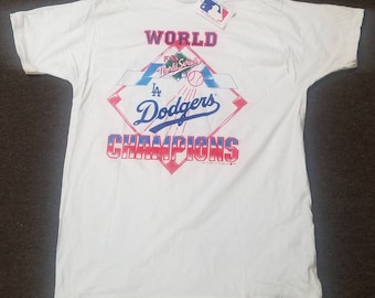 Puede incluir: Camiseta blanca con un estampado gráfico azul y rojo que dice "WORLD 1988 World Series LA Dodgers CHAMPIONS".
