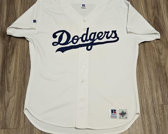 May include: White baseball jersey with the word "Dodgers" in navy blue script across the chest. The jersey has a v-neck, short sleeves, and a button-up front. A small "R" logo is on the left sleeve. A Russell Athletic tag is on the bottom.