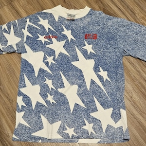 May include: A short-sleeve t-shirt featuring a blue and white speckled star pattern. The shirt includes a red Adidas logo on the left chest and a red USA graphic on the right chest. The design covers the front of the garment with a crew neckline.