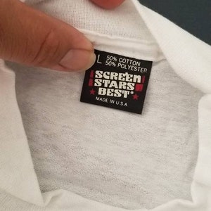 Op de afbeelding: Een wit T-shirt met een zwart label waarop staat "Screen Stars Best", "50% Katoen 50% Polyester" en "Made in U.S.A."