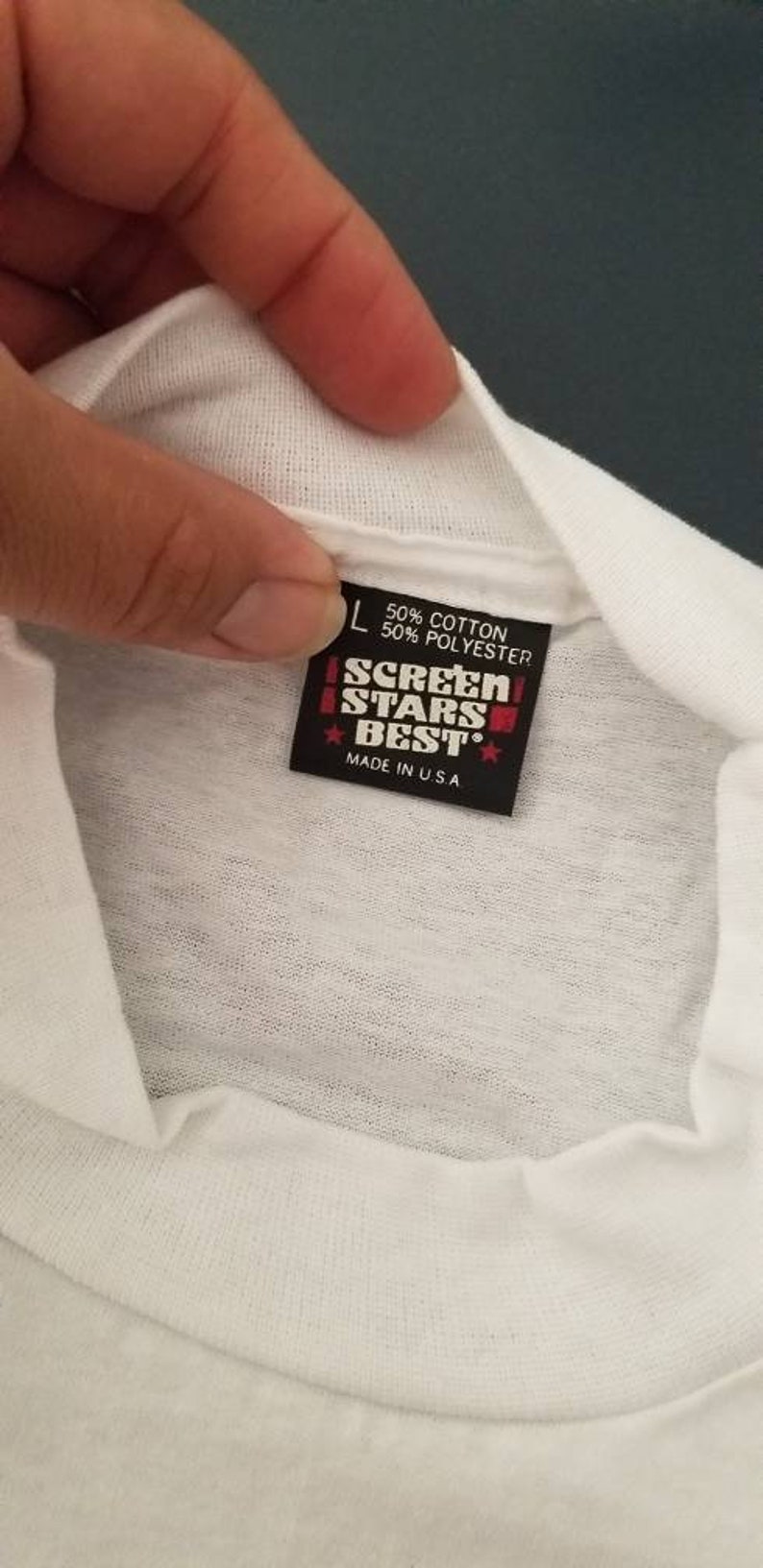 Op de afbeelding: Een wit T-shirt met een zwart label waarop staat "Screen Stars Best", "50% Katoen 50% Polyester" en "Made in U.S.A."
