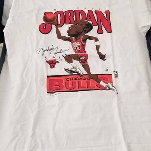 Op de afbeelding: Wit T-shirt met een cartoon afbeelding van Michael Jordan die basketbal speelt voor de Chicago Bulls. De tekst "MICHAEL JORDAN" is in rode letters boven de afbeelding gedrukt. De tekst "CHICAGO BULLS" is in rode letters onder de afbeelding gedrukt.