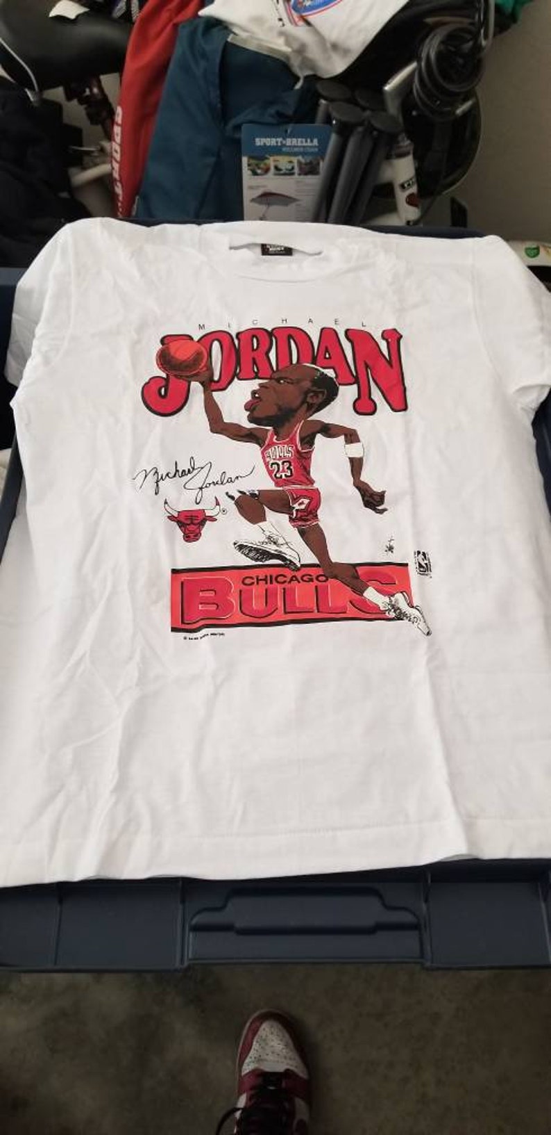 Op de afbeelding: Wit T-shirt met een cartoon afbeelding van Michael Jordan die basketbal speelt voor de Chicago Bulls. De tekst "MICHAEL JORDAN" is in rode letters boven de afbeelding gedrukt. De tekst "CHICAGO BULLS" is in rode letters onder de afbeelding gedrukt.