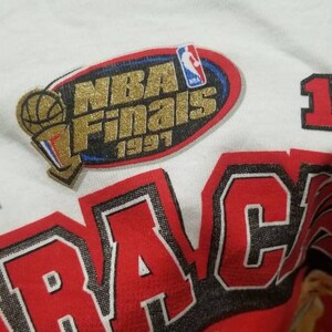May include: White t-shirt with red and black lettering that says "1992" and "TRACAMP" with a gold and blue patch that says "NBA Finals 1997".