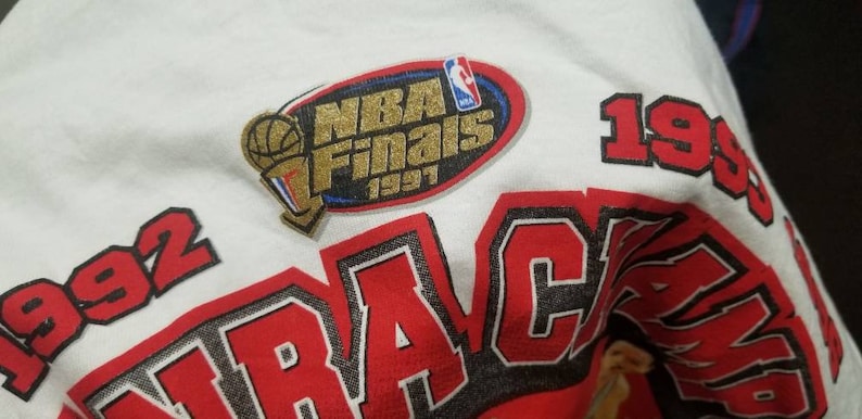 May include: White t-shirt with red and black lettering that says "1992" and "TRACAMP" with a gold and blue patch that says "NBA Finals 1997".