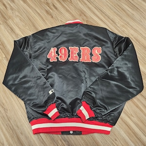 May include: Black satin bomber jacket with red and white striped cuffs and waistband. The back of the jacket features a large red and white "49ERS" logo.