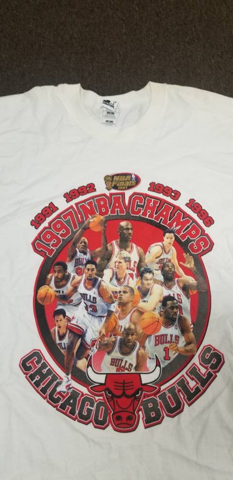 May include: White t-shirt with a red, black, and white graphic of the Chicago Bulls NBA Champions from 1991, 1992, 1993, and 1996. The graphic features a red bull mascot and a group of basketball players, including Michael Jordan.