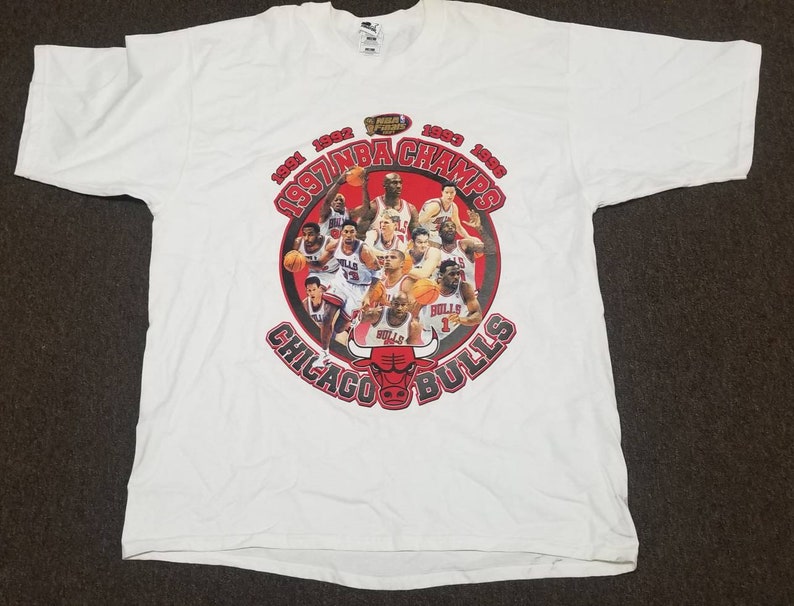 May include: White t-shirt with a graphic of the Chicago Bulls basketball team. The graphic features the team's logo and the text "1991 1992 1993 1996 1997 NBA CHAMPS Chicago Bulls".