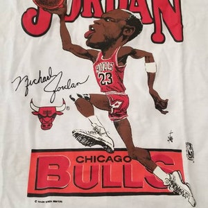 Op de afbeelding: Wit T-shirt met een rode en zwarte cartoon afbeelding van Michael Jordan die basketbal speelt. De tekst "MICHAEL JORDAN" staat in rode letters boven de afbeelding. De tekst "CHICAGO BULLS" staat in rode letters onder de afbeelding. De handtekening "Michael Jordan" staat in zwarte inkt onder de afbeelding.
