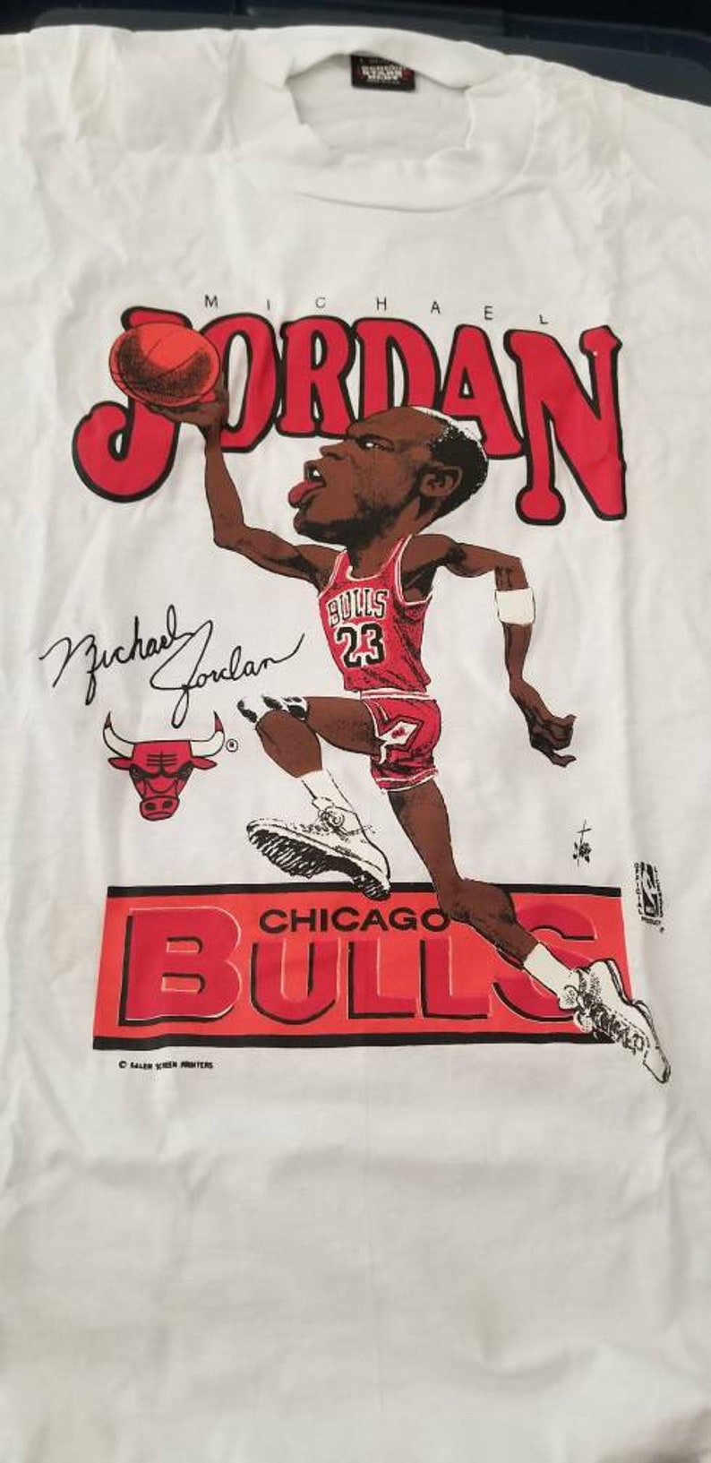 Op de afbeelding: Wit T-shirt met een rode en zwarte cartoon afbeelding van Michael Jordan die basketbal speelt. De tekst "MICHAEL JORDAN" staat in rode letters boven de afbeelding. De tekst "CHICAGO BULLS" staat in rode letters onder de afbeelding. De handtekening "Michael Jordan" staat in zwarte inkt onder de afbeelding.