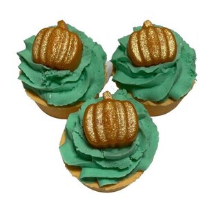 May include: Three cupcakes with green frosting and a gold-colored pumpkin decoration on top. The cupcakes sit on a light brown base, and the background is white. The cupcakes are a festive treat.
