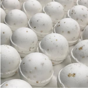 May include: A collection of white bath bombs with a spherical shape and a textured surface. Each bath bomb has a light dusting of brown specks. The bath bombs are arranged closely together, filling the frame.