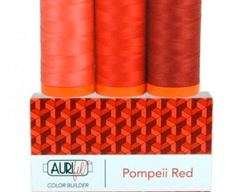May include: Three spools of red thread in varying shades, from light coral to deep crimson, sit atop a box with a geometric pattern. The box features the text "Pompeii Red" and the brand name "Aurifil Color Builder."