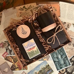 Puede incluir: Una caja de regalo llena de papel de embalaje marr&oacute;n y una variedad de art&iacute;culos, que incluyen un diario negro con un dise&ntilde;o de helecho de madera, una taza moteada en blanco y negro, una peque&ntilde;a bolsa marr&oacute;n, un juego de cartas con una escena de monta&ntilde;a y el texto "Outdoor Adventures Playing Cards", y una tela gris envuelta con cuerda.