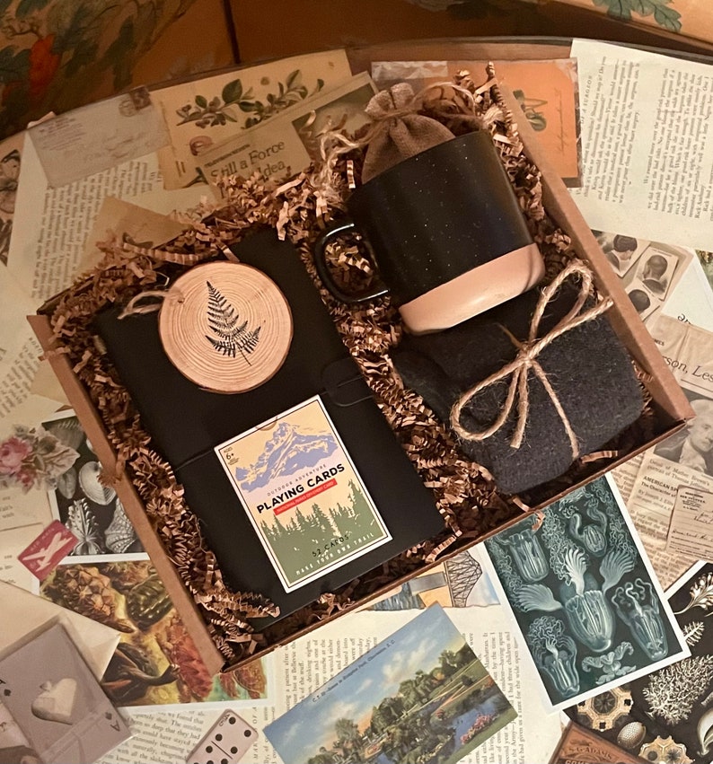 Puede incluir: Una caja de regalo llena de papel de embalaje marr&oacute;n y una variedad de art&iacute;culos, que incluyen un diario negro con un dise&ntilde;o de helecho de madera, una taza moteada en blanco y negro, una peque&ntilde;a bolsa marr&oacute;n, un juego de cartas con una escena de monta&ntilde;a y el texto "Outdoor Adventures Playing Cards", y una tela gris envuelta con cuerda.