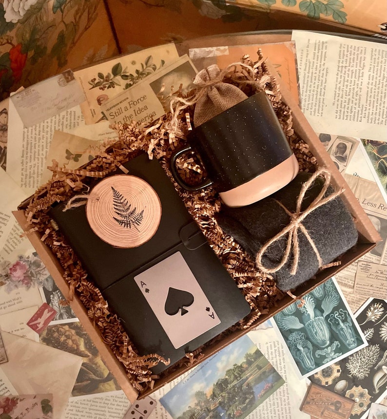 Puede incluir: Una caja de regalo llena de una taza negra y rosa, una peque&ntilde;a bolsa marr&oacute;n, un diario negro con un dise&ntilde;o de helecho, una carta de juego negra con el as de picas y una bufanda gris atada con cuerda.