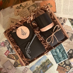 Puede incluir: Una caja de regalo llena de un diario negro, una taza negra y blanca, un peque&ntilde;o adorno de madera de helecho, una pistola de juguete negra y plateada y una bufanda de tela marr&oacute;n. La caja est&aacute; llena de papel arrugado marr&oacute;n.