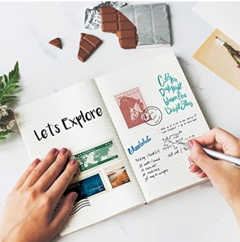Puede incluir: Un cuaderno abierto con las palabras "Let's Explore" escritas en la p&aacute;gina izquierda. La p&aacute;gina derecha tiene una lista titulada "Wanderlust" con una lista de verificaci&oacute;n de empaque. Hay varios sellos postales en la p&aacute;gina izquierda.