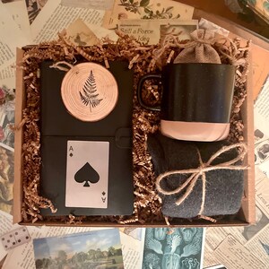 Puede incluir: Una caja de regalo llena de un diario negro con un dise&ntilde;o de helecho, una taza negra y beige, un par de calcetines gris oscuro y una carta de juego con el as de picas.