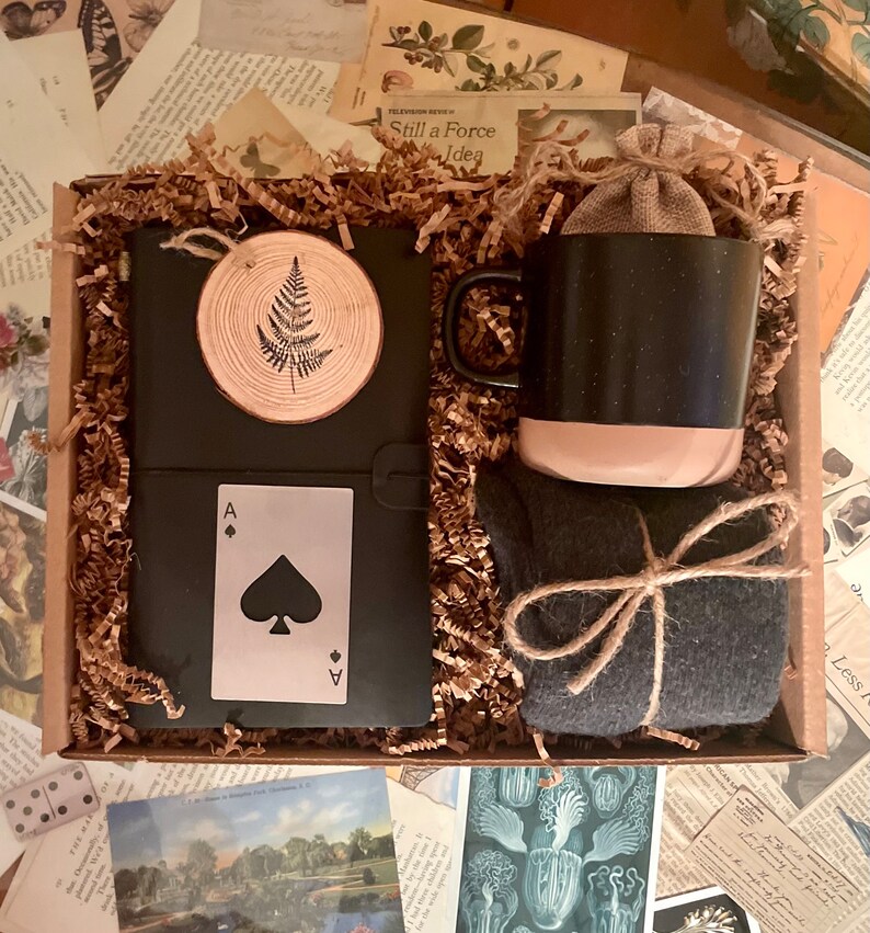 Puede incluir: Una caja de regalo llena de un diario negro con un dise&ntilde;o de helecho, una taza negra y beige, un par de calcetines gris oscuro y una carta de juego con el as de picas.