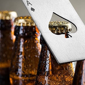 Puede incluir: Un abridor de botellas plateado con forma de as de picas. El abridor tiene una forma de pica recortada para abrir botellas. El abridor est&aacute; sobre cuatro botellas de vidrio marr&oacute;n.