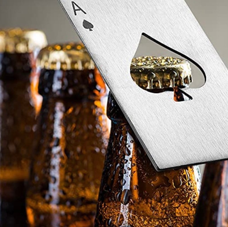 Puede incluir: Un abridor de botellas plateado con forma de as de picas. El abridor tiene una forma de pica recortada para abrir botellas. El abridor est&aacute; sobre cuatro botellas de vidrio marr&oacute;n.