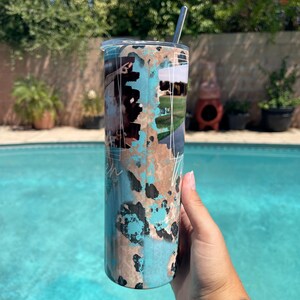 K&ouml;nnte beinhalten: Ein Edelstahlbecher mit Deckel und Strohhalm. Der Becher hat ein t&uuml;rkisfarbenes, braunes und schwarzes Kuhfellmuster mit wei&szlig;er Schrift. Der Becher wird von einer Person in der N&auml;he eines Swimmingpools gehalten.