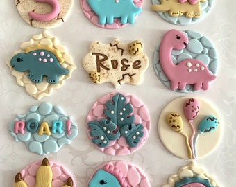 May include: Twelve cupcake toppers featuring a dinosaur theme. The toppers are made of fondant and feature various dinosaurs, including a blue dinosaur, a pink dinosaur, a yellow dinosaur, and a green dinosaur. The toppers also feature a footprint, leaves, balloons, and the words "Roar!" and "Rose".