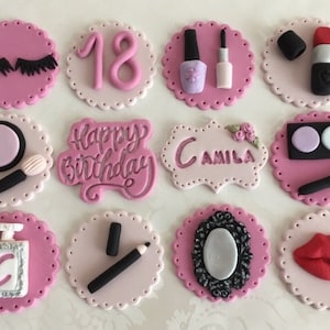 May include: Twelve pink and white cupcake toppers with various makeup-themed designs, including eyelashes, lipstick, nail polish, a compact, a brush, a mirror, and the words "Happy Birthday" and "Camila".