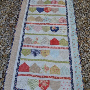 May include: A white quilt with a patchwork design featuring various floral patterns, red and blue accents, and a scalloped border. The quilt is made up of rows of houses and hearts.