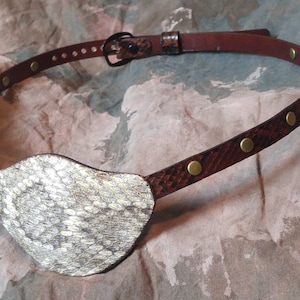 May include: A brown leather eye patch with a snakeskin design and brass accents. The eye patch is attached to a brown leather strap with brass studs.