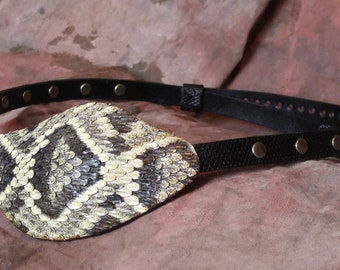 Könnte beinhalten: Ein schwarzes Leder-Augenpflaster mit Schlangenhaut-Design. Das Pflaster hat ein Band mit silbernen Nieten.