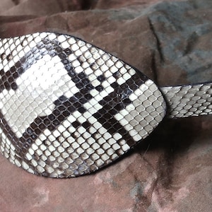 May include: A black and white snakeskin eye patch with a strap. The eye patch is made of real snakeskin and has a unique pattern.