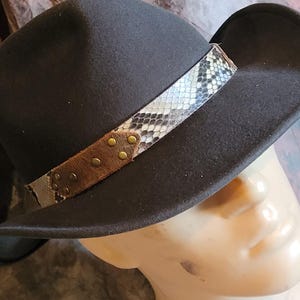 May include: A black felt fedora hat with a snakeskin patterned band and brown leather accents. The band has gold-coloured metal studs.