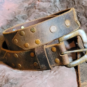 May include: A brown leather belt with a silver buckle and brass studs. The belt is worn and has a distressed look.