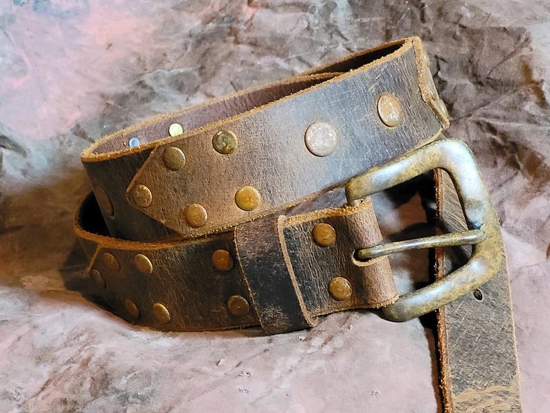 May include: A brown leather belt with a silver buckle and brass studs. The belt is worn and has a distressed look.