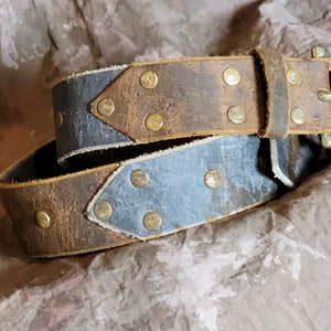 May include: A brown leather belt with a blue leather accent and brass rivets. The belt has a simple buckle.