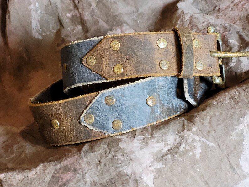 May include: A brown leather belt with a blue leather accent and brass rivets. The belt has a simple buckle.