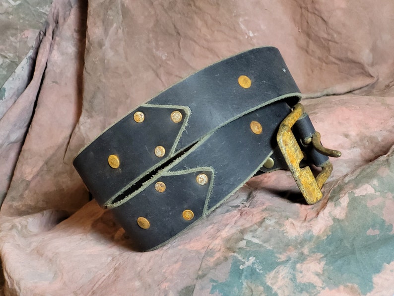 May include: A black leather belt with a brass buckle and gold-toned rivets. The belt is folded in half and the buckle is visible.