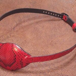May include: A red leather eye patch with a snakeskin pattern. The patch is attached to a black leather strap with adjustable holes.