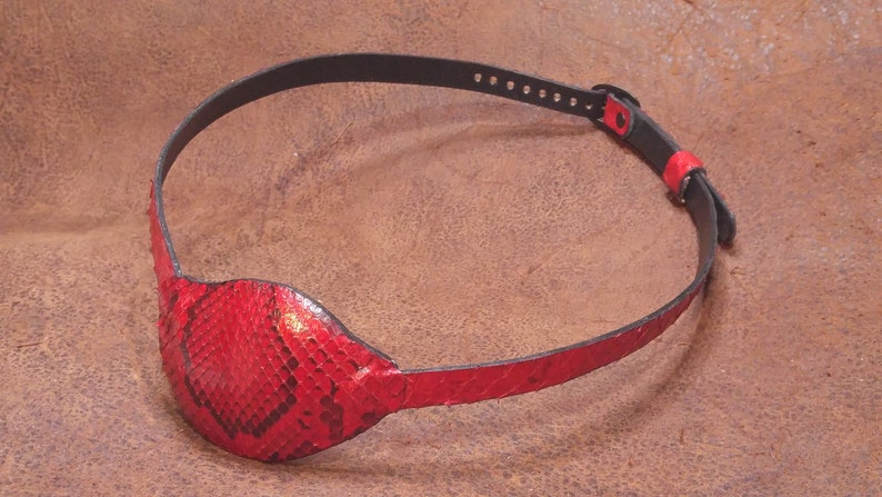 May include: A red leather eye patch with a snakeskin pattern. The patch is attached to a black leather strap with adjustable holes.