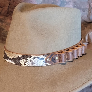 May include: A tan felt fedora hat featuring a decorative band. The band is composed of a section of snakeskin pattern material and a brown leather strip with integrated loops, secured by small metallic rivets at the side of the hat crown.