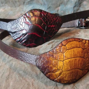 May include: Two leather eye patches with adjustable straps. One patch is a deep red and black, the other is a golden brown. Both have a textured, reptile-like pattern and brown leather straps with a buckle.