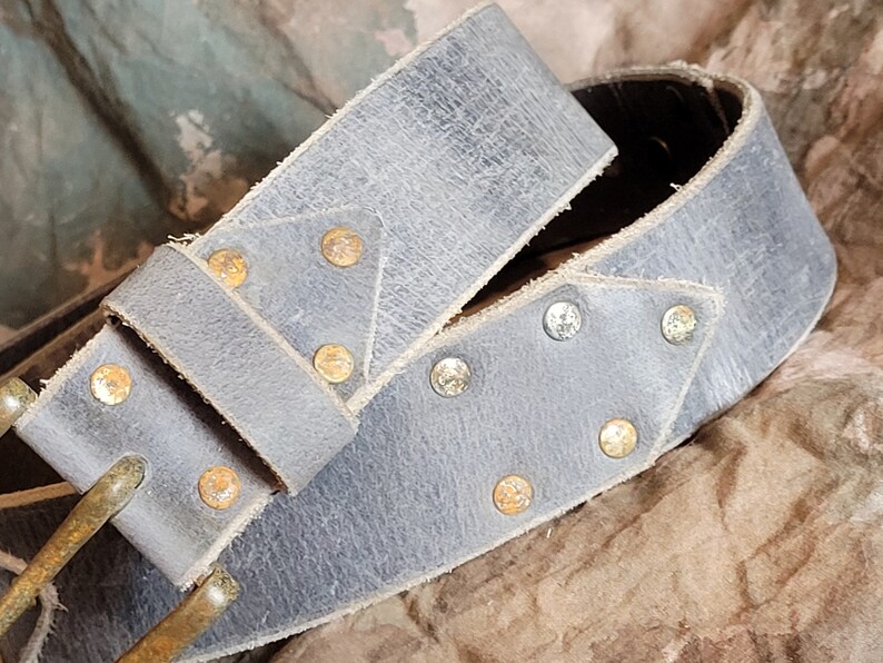 May include: A gray leather belt with a brass buckle and decorative metal studs. The belt is folded in half and the buckle is visible.