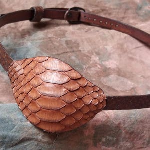 May include: Brown leather eye patch with a textured, scale-like design. The eye patch is attached to a brown leather strap with an adjustable buckle. The item is designed to be worn around the head.