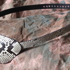 May include: A black leather eye patch with a white and black snakeskin design. The eye patch is attached to a black leather strap with holes for adjusting the fit.