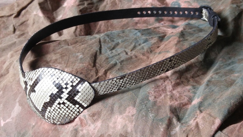 May include: A black leather eye patch with a white and black snakeskin design. The eye patch is attached to a black leather strap with holes for adjusting the fit.