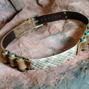 May include: A brown leather belt with a snakeskin pattern and a brass buckle. The belt has a brown leather ammo holder with six empty shell casings.
