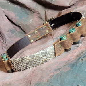 Peut inclure: Une ceinture en cuir marron avec un motif de peau de serpent et une rangée de douilles de balles en métal. La ceinture est fermée par une boucle en laiton.