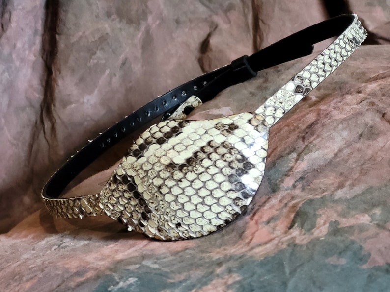 May include: A brown leather eye patch with a snakeskin design. The patch is attached to a black leather strap.